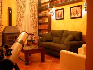 Hermosa biblioteca con paredes llenos de libros sobre estanterías de madera. Al fondo dos ventanales con cortinas beige y estampado verde oscuro a ambos lados de una chimenea de forja. Dos cómodos sofás beige junto a una mesa de centro permiten descansar. En la imagen se observa un telescopio para ver las estrellas.
