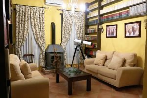 Hermosa biblioteca con paredes llenos de libros sobre estanterías de madera. Al fondo dos ventanales con cortinas beige y estampado verde oscuro a ambos lados de una chimenea de forja. Dos cómodos sofás junto a una mesa de centro permiten descansar.