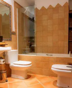 Baño de habitación con bañera, lavabo cuadrado como en tiempos antiguos, azulejos rústicos en tonos tostados y preciosa cenefa en tonos azules y grises, imitando la cerámica antigua andaluza.