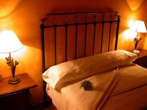 La cama de forja flanqueada por apliques rústicos está abierta invitando al viajero a dormir, con algunas flores frescas sobre la almohada.