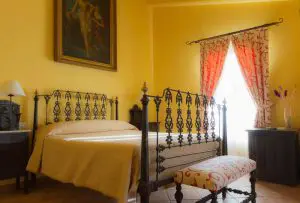 Señorial cama de forja de principios del siglo XX, con flores adornando sus patas. Sobre la cama un antiguo cuadro de temática mitológica, el baño de Diana. Al fondo un balcón con hermosas cortinas estampadas de beige y motivos burdeos.