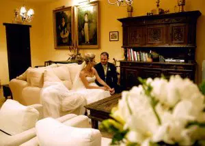 Detalle del acogedor salón con un gran sofá sobre el que se sientan una pareja de novios que se miran enamorados. Al fondo dos cuadros de un niño y una niña de estilo goyesco mejicano. El ramo de flores de la novia en primer plano, da frescura y color a la imagen.